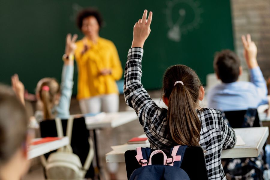 Rear view of schoolgirl raising her arm to answer the question in the classroom.