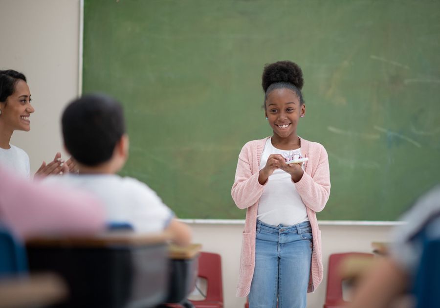 Girl stands in front of class to speak with teach clapping in the corner