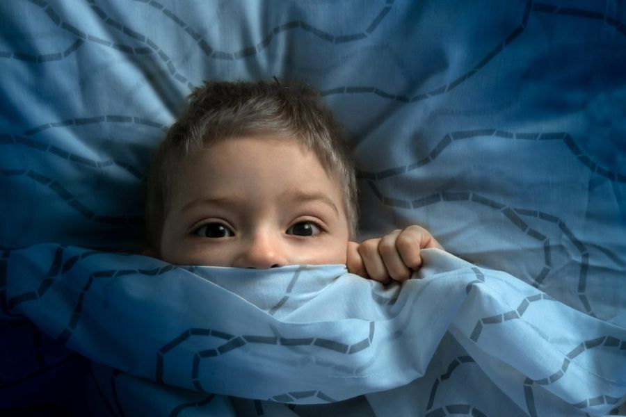 A boy pulls up his covers to his nose with his eyes wide open because he is scared of the dark and bad dreams in his bed.