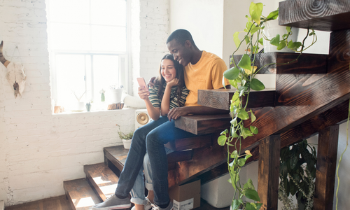 A couple sits on the stairs together smiling and looking at a cell phone